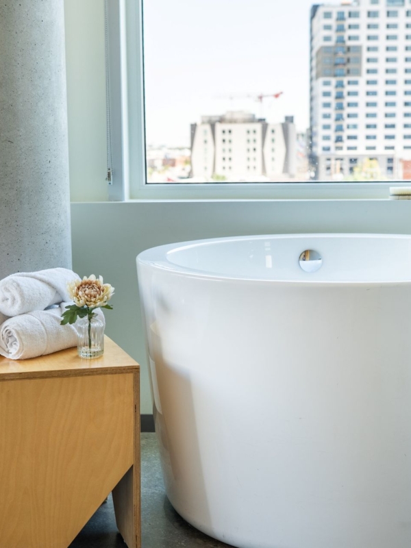 A white bathtub with a wooden nightstand and towels next to it, and a window showing a city view.