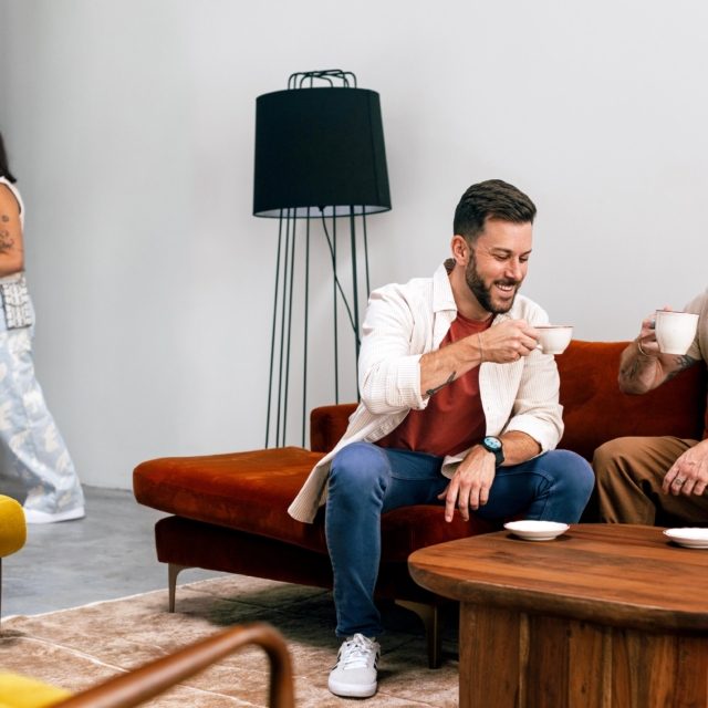 A man sits on a brown couch with two other people partially visible, holding a cup of coffee in a lobby setting.