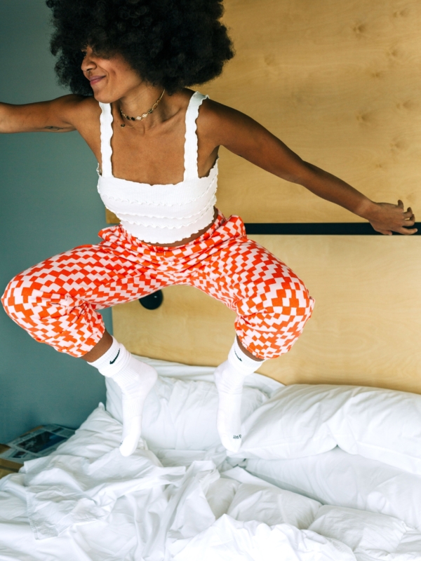 A woman in orange and white patterned pants jumping on a bed with white bedding.