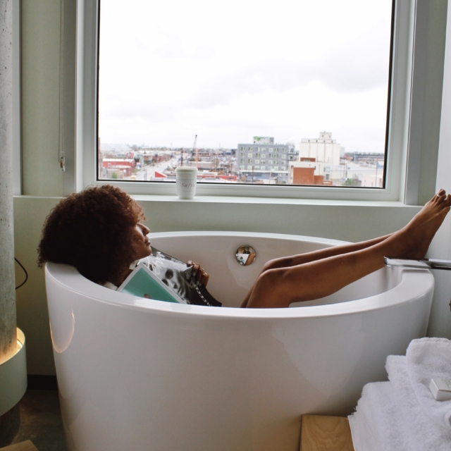 A woman relaxes in a freestanding bathtub in front of a large window with a city view.