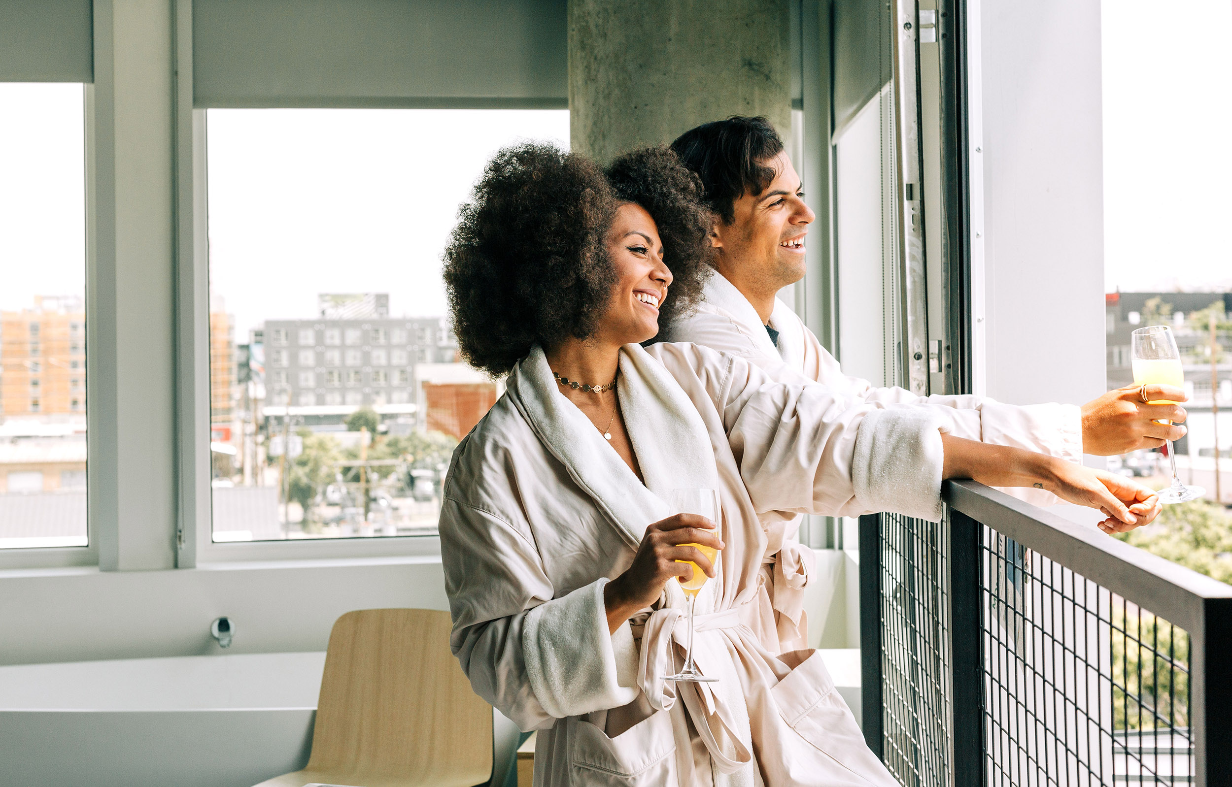 A couple in white robes stands on a balcony with a city view, holding champagne glasses.